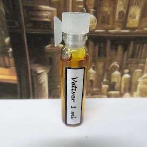 以下が含まれることがあります： 「Vetiver 1 ml」とラベルされた、金色の液体が入った小さなガラス瓶。白いキャップと黒い文字の白いラベルが付いています。背景はぼやけており、さまざまなアイテムが置かれた棚が見えます。