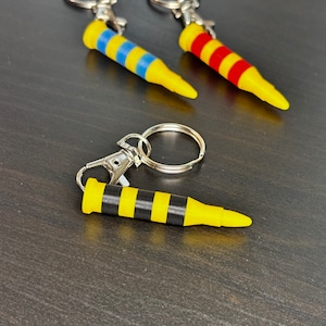 May include: Three keychains shaped like bullets with yellow and black stripes, red and yellow stripes, and blue and yellow stripes. The keychains are on a dark gray surface.