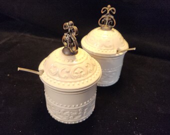 May include: Two white ceramic sugar bowls with ornate designs and metal lids. Each bowl has a small spoon attached to the lid.