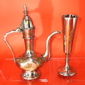 May include: A silver and gold teapot with a floral design and a matching silver and gold goblet. The teapot has a lid and a spout. The goblet has a wide base and a narrow top.