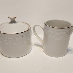May include: A white ceramic sugar bowl with a lid and a creamer. Both pieces have a gold trim and embossed floral designs. The sugar bowl is round, and the creamer has a handle and spout. The set is on a white surface.