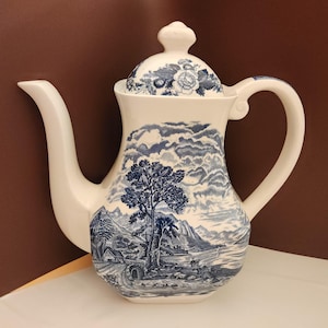 May include: A white ceramic teapot with a blue and white floral design on the lid and a blue and white landscape scene on the body. The teapot has a spout and a handle.