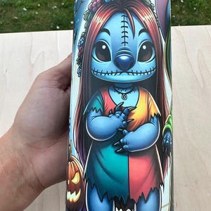 May include: A cylindrical tumbler featuring a cartoon character in a Halloween theme. The character has blue skin, long red hair, and is wearing a patchwork dress. The design includes a jack-o'-lantern and a night sky background.
