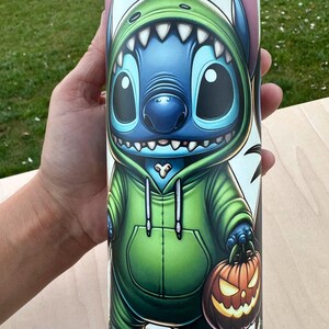 May include: A cylindrical tumbler featuring a cartoon character in a green hoodie and holding a jack-o'-lantern. The character is blue with large eyes and pink ears. The tumbler has a white background with black bat silhouettes.