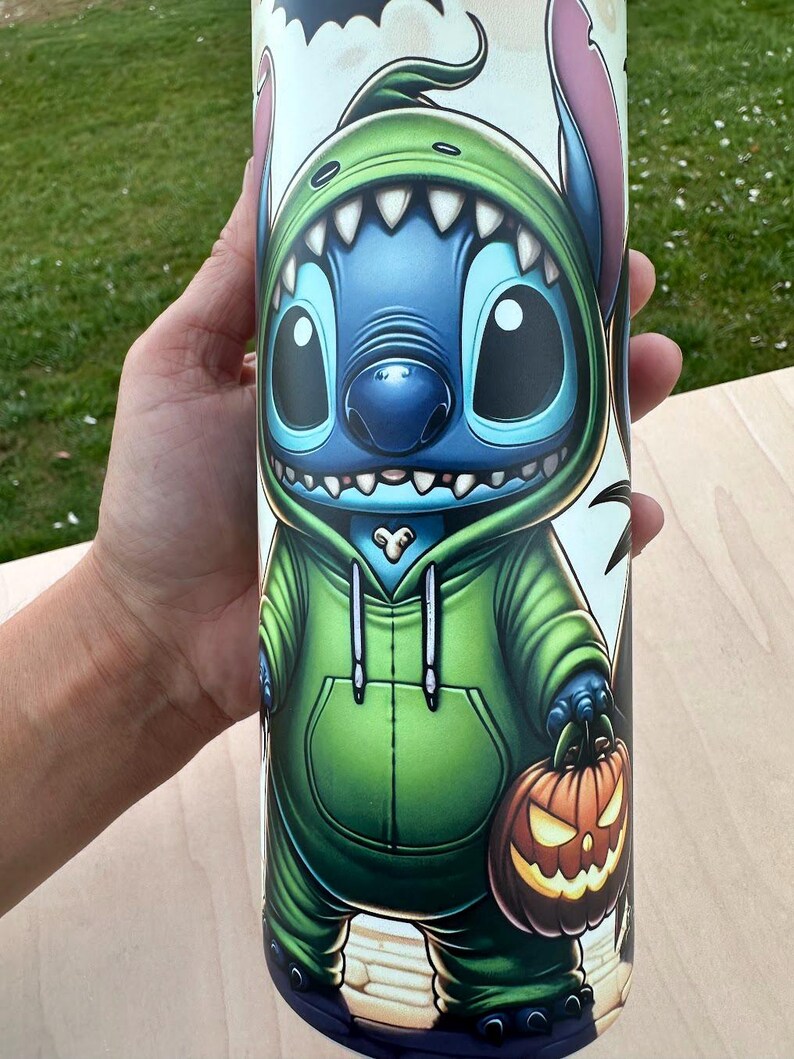 May include: A cylindrical tumbler featuring a cartoon character in a green hoodie and holding a jack-o'-lantern. The character is blue with large eyes and pink ears. The tumbler has a white background with black bat silhouettes.