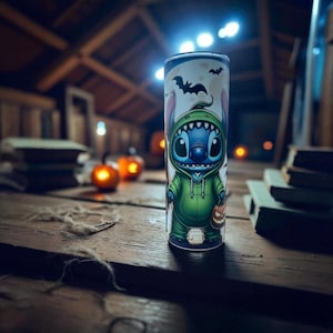 May include: A Halloween-themed tumbler featuring the character Stitch in a green hoodie, holding a jack-o'-lantern. The tumbler has a white background with bats and a moon. The design is printed on a cylindrical, insulated tumbler.