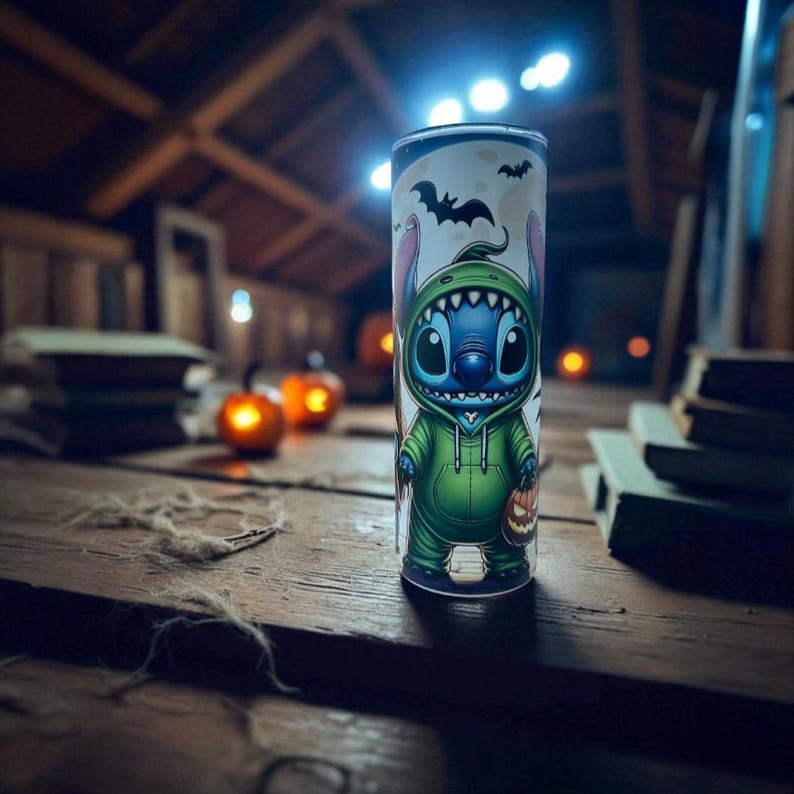 May include: A Halloween-themed tumbler featuring the character Stitch in a green hoodie, holding a jack-o'-lantern. The tumbler has a white background with bats and a moon. The design is printed on a cylindrical, insulated tumbler.