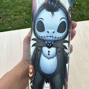 May include: A tall, cylindrical tumbler featuring a cartoon illustration of Jack Skellington from The Nightmare Before Christmas. The design includes a black and white pinstripe suit, a bat, and a pumpkin. The tumbler is held by a person.