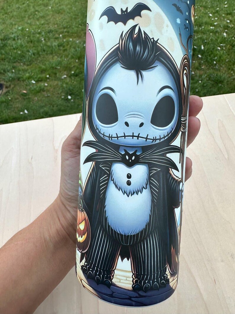May include: A tall, cylindrical tumbler featuring a cartoon illustration of Jack Skellington from The Nightmare Before Christmas. The design includes a black and white pinstripe suit, a bat, and a pumpkin. The tumbler is held by a person.