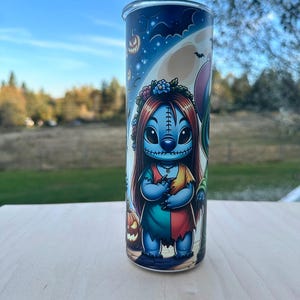 May include: A tall, cylindrical tumbler featuring a Halloween-themed design. The artwork depicts a blue character with red hair and a floral crown, dressed in a patchwork dress. The background includes a full moon, bats, and jack-o'-lanterns, set against a blue sky.
