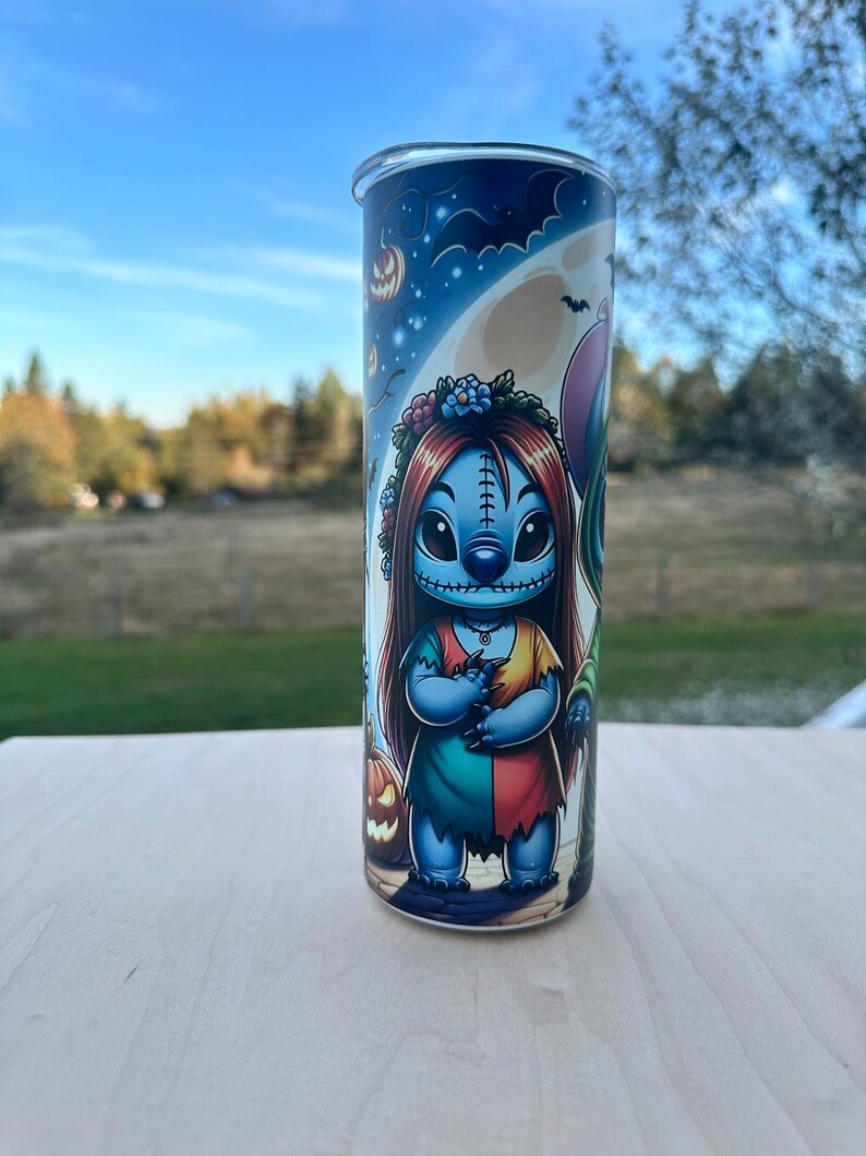 May include: A tall, cylindrical tumbler featuring a Halloween-themed design. The artwork depicts a blue character with red hair and a floral crown, dressed in a patchwork dress. The background includes a full moon, bats, and jack-o'-lanterns, set against a blue sky.