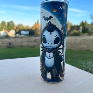 May include: A tall, cylindrical tumbler featuring a cartoon character in a black suit with a white shirt and a bat. The tumbler has a blue background with pumpkins and a full moon. The tumbler is made of stainless steel.