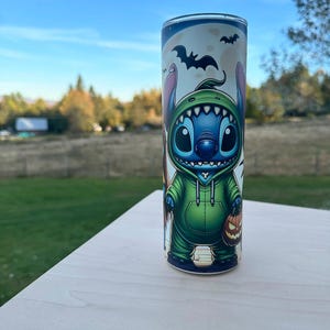 May include: A cylindrical tumbler featuring a cartoon character in a green hoodie, holding a jack-o'-lantern. The design includes bats and a full moon, with a blue and white background. The tumbler is set on a light-colored surface.