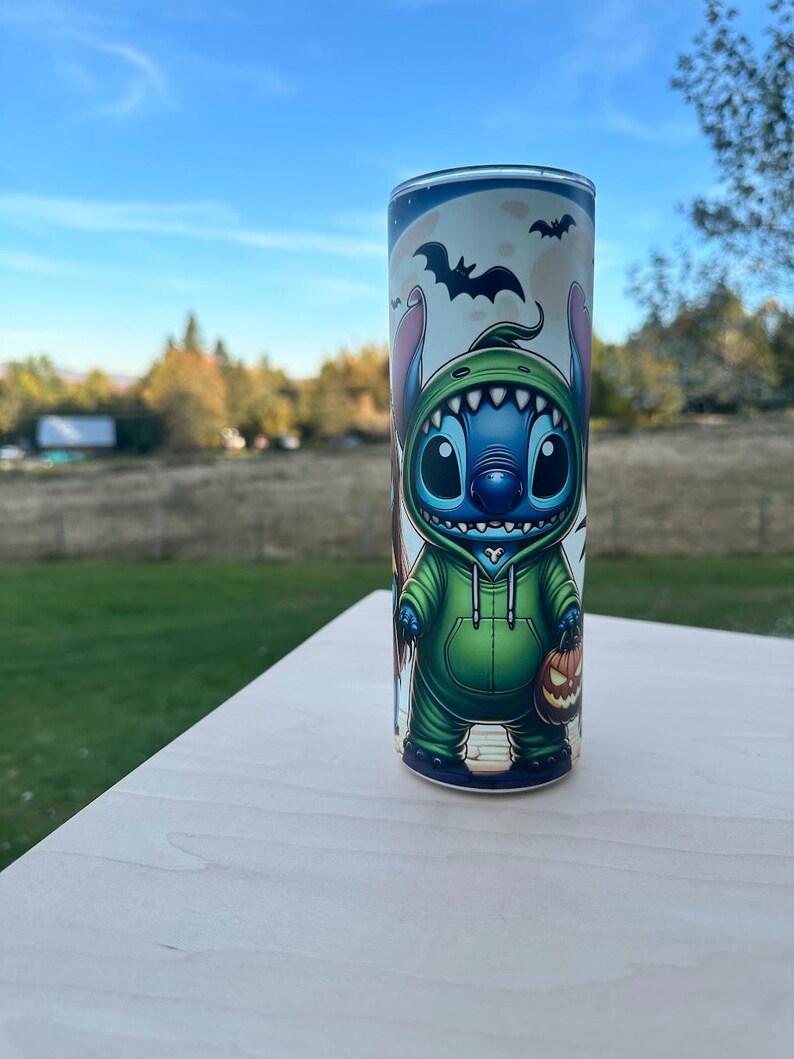 May include: A cylindrical tumbler featuring a cartoon character in a green hoodie, holding a jack-o'-lantern. The design includes bats and a full moon, with a blue and white background. The tumbler is set on a light-colored surface.