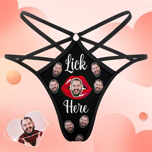 Puede incluir: Braga tanga negra con un dise&ntilde;o &uacute;nico. El panel frontal presenta el texto "Lick Here" sobre un gr&aacute;fico de labios rojos con una cara en el centro. M&uacute;ltiples caras rodean el gr&aacute;fico de labios. Las correas son negras.