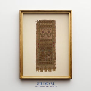 May include: A framed art print featuring a vertical textile fragment with brown, tan, and muted red tones. The design includes symmetrical floral motifs and two stylized figures at the base, finished with a row of fringe at the bottom edge.