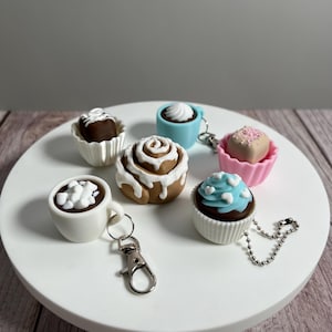 以下が含まれることがあります： シナモンロール、カップケーキ、ホットチョコレートのカップなど、食べ物をテーマにしたミニチュアキーホルダーのコレクション。キーホルダーは、茶色、白、青などのさまざまな色で、白い円形の表面に配置されています。