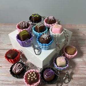 Pode incluir: Uma coleção de chaveiros de trufas de chocolate em miniatura em várias cores. Cada trufa está aninhada em uma xícara colorida e canelada e presa a um chaveiro prateado. As trufas são decoradas com granulado, redemoinhos e outros desenhos.