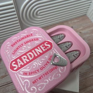 Op de afbeelding: Een roze rechthoekige houder met een deksel voorzien van de tekst SUSTAINABLE LIGHTLY SMOKED SARDINES IN SPICY OLIVE OIL NET WT 7 OZ (200g) en een zilveren lipje. Binnenin liggen drie kleine zilveren visfiguren met zichtbare ogen.