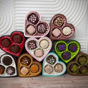 May include: An assortment of heart-shaped boxes filled with chocolates. The boxes are in various colors, including red, black, pink, and green. Each box contains several chocolates with different toppings and designs, such as sprinkles and swirls.