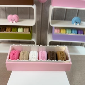 May include: A collection of miniature macaron sets displayed on white tiered shelves. Each set features colorful, textured macaron cookies arranged in rectangular boxes. A single pink macaron charm hangs above, and small heart-shaped figures are included.