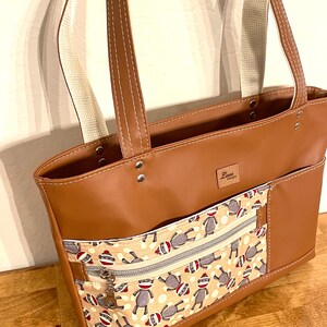 May include: A brown leather tote bag with a zippered pocket and a monkey print lining. The bag has two straps and a label that says "Zane Designs".