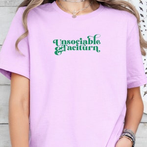 Puede incluir: Una camiseta lavanda con el texto "Unsociable & Taciturn" en verde.
