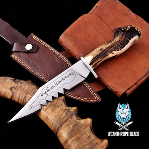 May include: A handcrafted hunting knife with a serrated blade and a handle made from deer antler. The knife features an engraved design on the blade and comes with a brown leather sheath. The knife is displayed on a wooden surface.