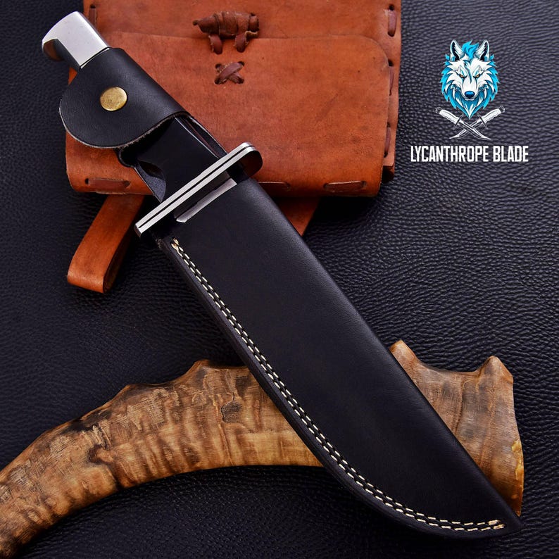 Puede incluir: Una funda de cuchillo de cuero negro con costuras blancas. La funda est&aacute; cerrada y tiene un clip de metal en la parte superior. La funda est&aacute; sobre una superficie de madera marr&oacute;n.