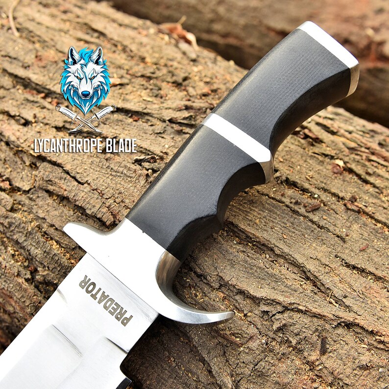 May include: A black and silver hunting knife with a black handle and a silver blade. The blade has the word "PREDATOR" engraved on it. The knife is resting on a piece of wood.