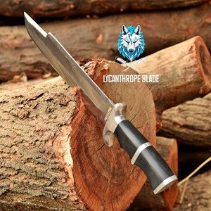 May include: A silver knife with a black handle and the word "PREDATOR" on the blade. The knife is resting on a wooden log. The background is blurred and shows more logs.