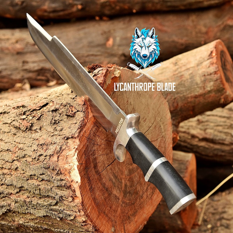 May include: A silver knife with a black handle and the word "PREDATOR" on the blade. The knife is resting on a wooden log. The background is blurred and shows more logs.