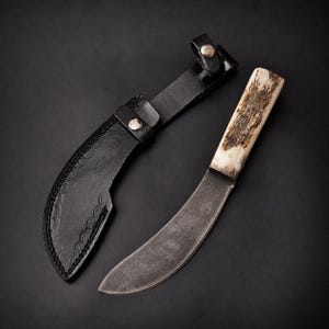 Könnte beinhalten: Ein feststehendes Messer mit einer gebogenen Stahlklinge und einem strukturierten Geweihgriff, begleitet von einer schwarzen Lederscheide mit dekorativen Bogenkanten-Nähten und einem Metalldruckknopf an der Gürtelschlaufe.