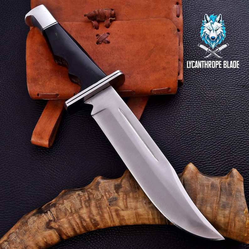 Puede incluir: Un cuchillo de caza plateado y negro con mango negro y funda de cuero. El cuchillo est&aacute; sobre una superficie de cuero marr&oacute;n con un logotipo de lobo y el texto "Lycanthrope Blade".