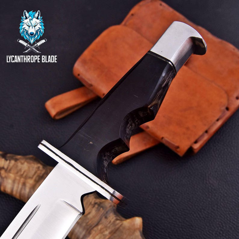 Puede incluir: Un cuchillo de caza negro y plateado con mango de madera y funda de cuero. El cuchillo tiene una hoja afilada y una punta puntiaguda. La funda es de cuero marr&oacute;n con costuras. El cuchillo se llama "Lycanthrope Blade".