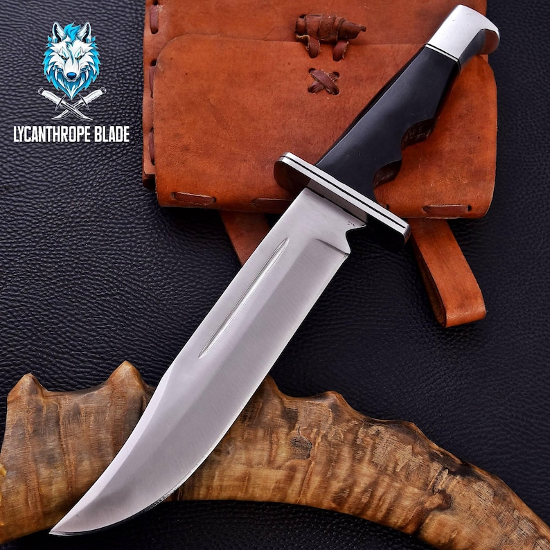 Puede incluir: Un cuchillo de caza grande, de color plateado, con mango negro y funda de cuero. El cuchillo est&aacute; apoyado en una pieza de madera con un patr&oacute;n marr&oacute;n y blanco.