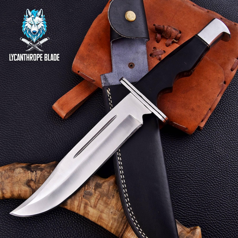 Puede incluir: Un cuchillo de caza con mango negro, hoja plateada y funda de cuero. El cuchillo est&aacute; sobre una superficie de cuero marr&oacute;n con el texto "LYCANTHROPE BLADE" en la esquina superior izquierda.