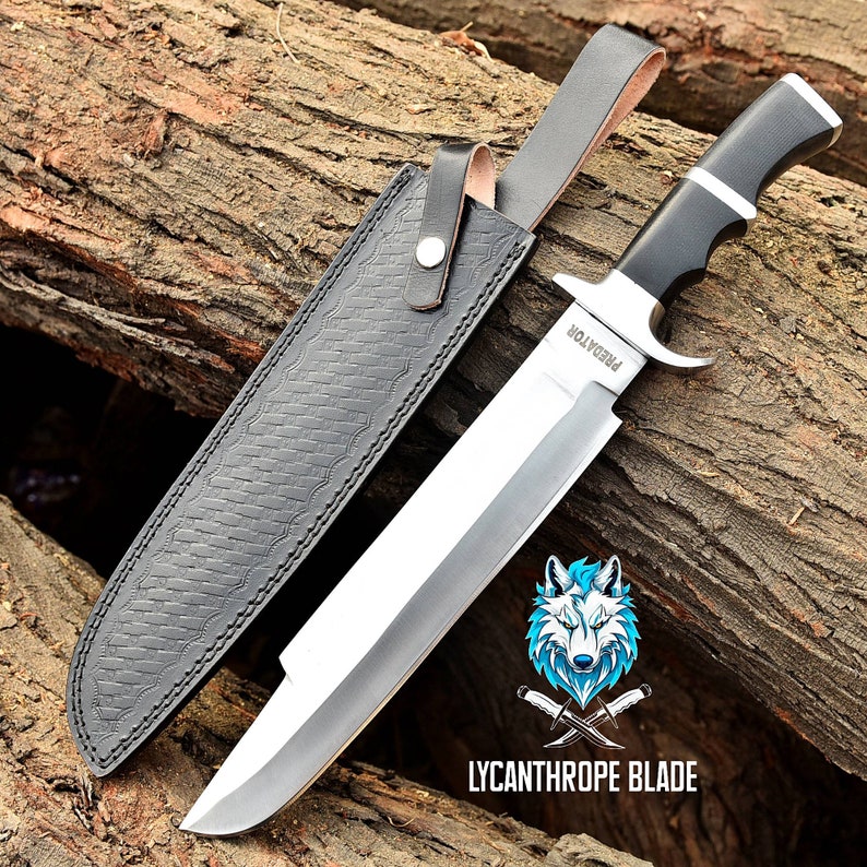 May include: A large, silver, fixed blade knife with a black handle and a leather sheath. The knife has the word "Predator" engraved on the blade. The knife is resting on a piece of wood.