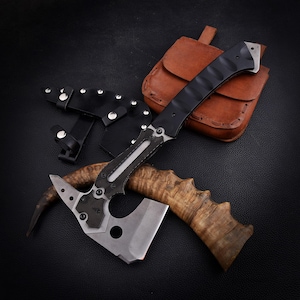 Pode incluir: Um machado tático com cabo preto e lâmina prateada, acompanhado por uma bainha de couro preta e uma bolsa de couro marrom. O machado apresenta um design único com uma lâmina curva e um cabo de madeira.
