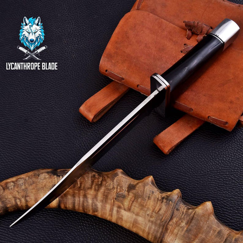 Puede incluir: Un cuchillo con mango negro, hoja plateada y pomo plateado. El cuchillo est&aacute; apoyado en una pieza de madera con una funda de cuero marr&oacute;n en el fondo. El texto "LYCANTHROPE BLADE" es visible en la imagen.
