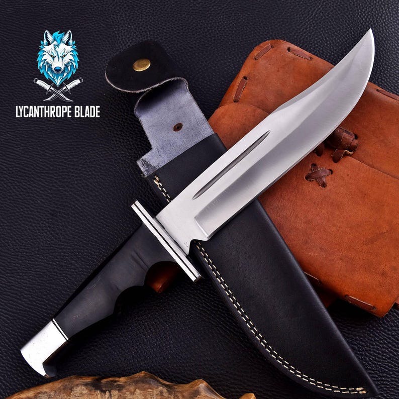 Puede incluir: Un cuchillo de caza con mango negro, hoja plateada y funda de cuero. La funda tiene una solapa de cuero marr&oacute;n con costuras decorativas.