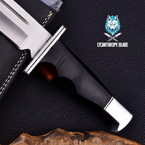 Puede incluir: Primer plano de un cuchillo de caza negro y plateado con mango de madera. El cuchillo est&aacute; sobre un fondo de cuero negro con el texto "LYCANTHROPE BLADE" en blanco.