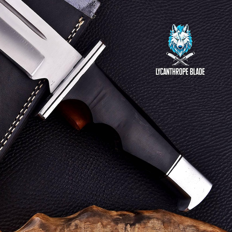 Puede incluir: Primer plano de un cuchillo de caza negro y plateado con mango de madera. El cuchillo est&aacute; sobre un fondo de cuero negro con el texto "LYCANTHROPE BLADE" en blanco.