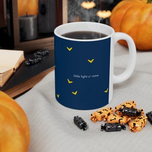 May include: A blue ceramic mug with a white handle and yellow stars. The mug has the text "little light o' mine" printed on it. The mug is filled with coffee and is sitting on a gray surface with orange and black candy.