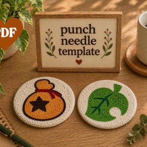 Puede incluir: Dos posavasos redondos de color blanquecino con diseños bordados. Uno presenta una bolsa de dinero con una estrella negra, el otro una hoja verde. Un letrero de madera dice "punch needle template". Una taza de café está cerca.