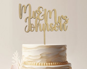 May include: A two-tiered white wedding cake with gold accents and pink flowers. The cake is topped with a wooden cake topper that says "Mr & Mrs Johnson".