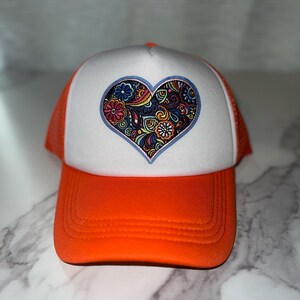 May include: Orange and white trucker hat with a colorful, patterned heart design on the front.