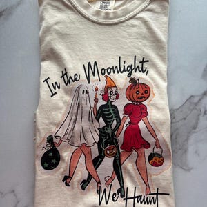 Peut inclure: Un t-shirt crème avec une illustration vintage de trois personnages en costumes d'Halloween. Le texte "In the Moonlight, We Haunt" est imprimé sur le t-shirt.