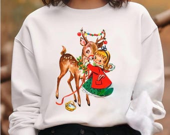 Op de afbeelding: Witte sweatshirt met een vintage kerstillustratie. Het ontwerp toont een hert met een slinger en een meisje in een rode jas. Het hert heeft een slinger van kleurrijke lichtjes op zijn gewei. Een mand en een rood lint staan ook in de afbeelding.