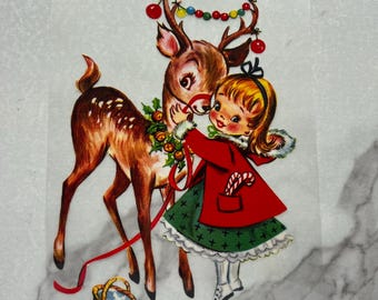 Op de afbeelding: Een vintage kerstkaart met een meisje in een rode jas en een groene rok die een snoepstok vasthoudt en een bruin en wit gevlekt hert omhelst met een rood lint en een krans van hulst om zijn nek. Het hert heeft een reeks kleurrijke kerstverlichting en rode ornamenten om zijn geweien.
